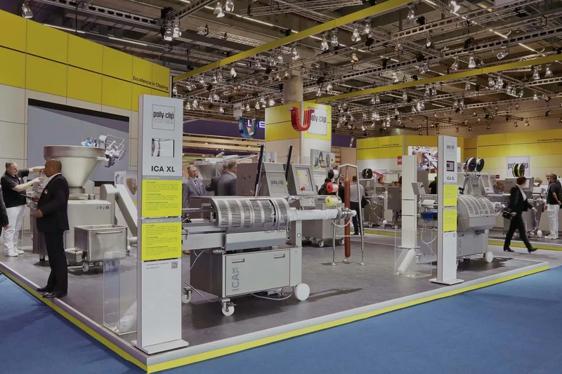 Suasana pameran mesin industri pengemasan dan pemrosesan makanan di hall pameran B2B dengan stan berlantai raised platform abu-abu, mesin clipping stainless steel, instalasi lighting truss gantung, dan interaksi profesional exhibitor