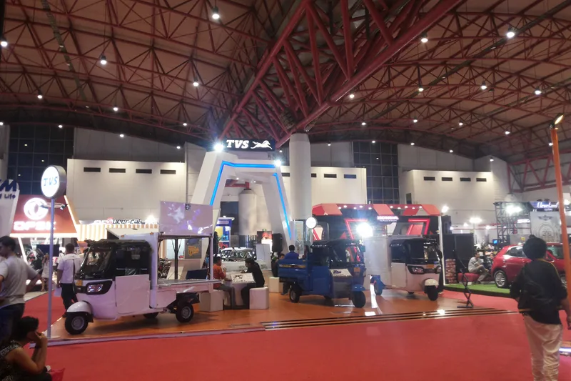 Suasana hall pameran otomotif indoor JIExpo Kemayoran dengan konstruksi rangka atap space-frame merah, booth display kendaraan niaga TVS, dan lorong karpet merah