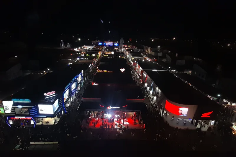 Pemandangan udara lorong pameran outdoor Jakarta Fair Kemayoran malam hari dengan paviliun pameran otomotif Yamaha dan Honda berlayar LED besar